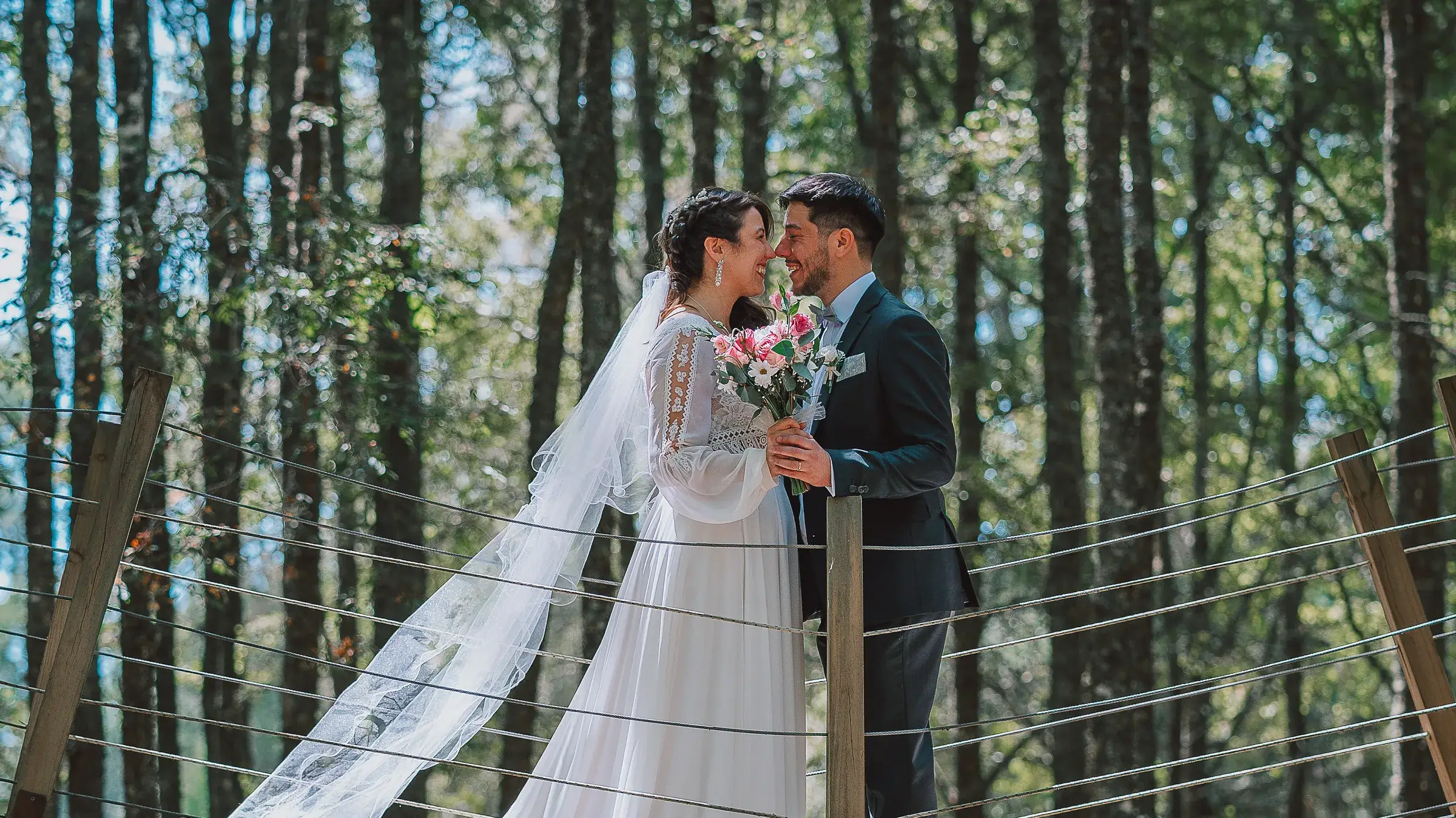 A bride and groom kiss on a rope bridge in a pine forest, her veil caught by the wind